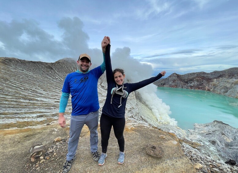 Two people standing on a rocky terrain with a geyser in the background. 2