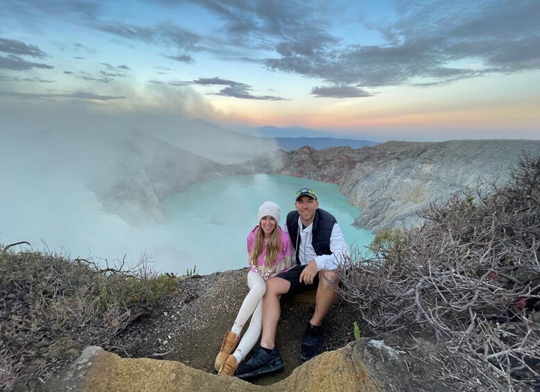 Two people sitting on a rocky outcrop overlooking a turquoise lake with steam rising from the water. 3