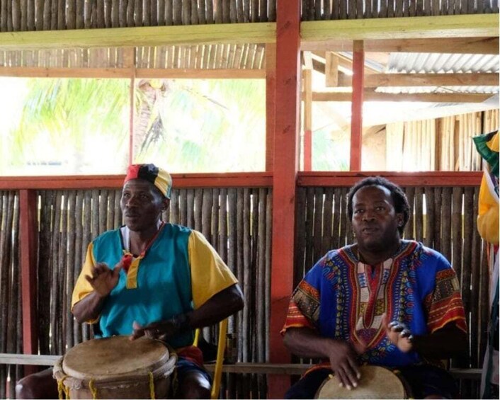 Two individuals playing drums in a rustic setting with bamboo walls and a corrugated metal roof. 2