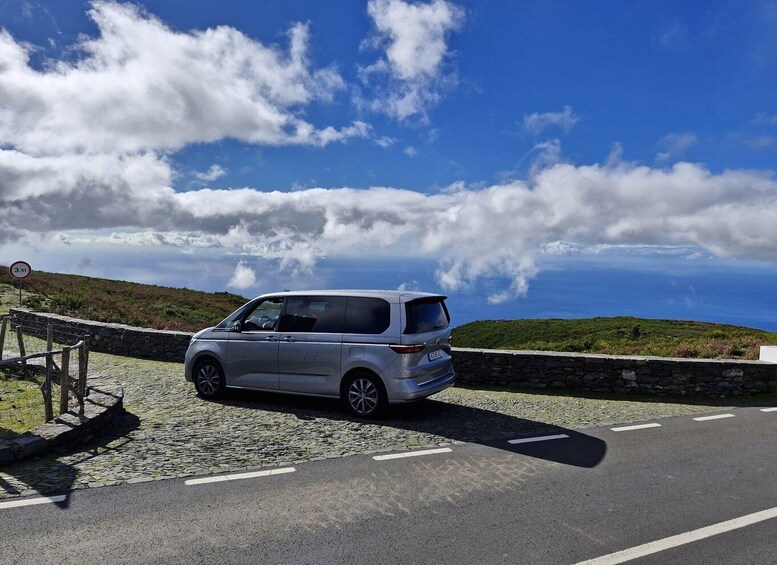 A silver SUV parked on a cobblestone road with a stone wall and a guardrail on the left, overlooking a coastal landscape with a clear blue sky and scattered clouds. 3