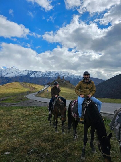 Two people riding horses on a road with a mountainous landscape in the background. 5