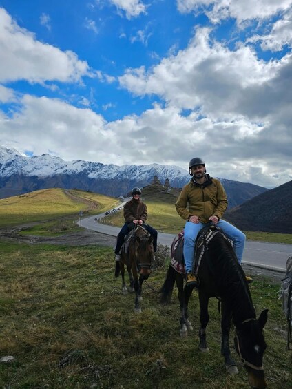 Two people riding horses on a road with a mountainous landscape in the background. 3