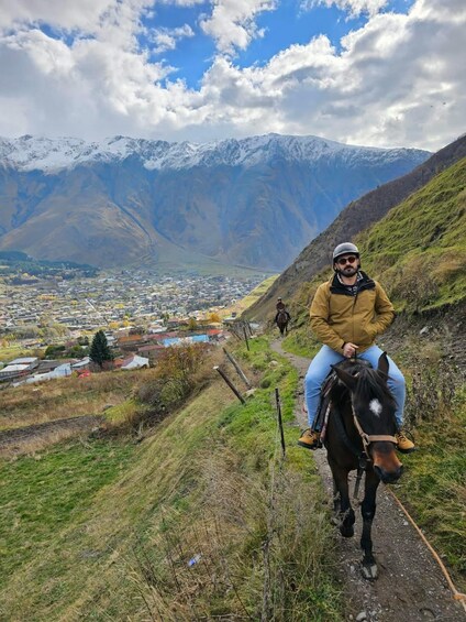 A person riding a horse on a mountain trail with a town in the valley below and snow-capped mountains in the distance. 4