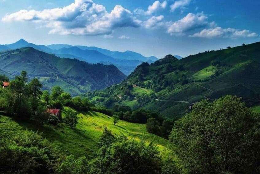 A lush green valley with rolling hills, a winding road, and distant mountains under a partly cloudy sky. 1