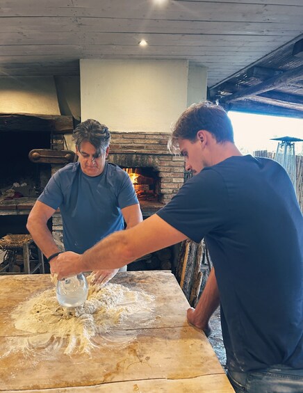 Two people kneading dough on a wooden table in a rustic kitchen with a brick fireplace. 1
