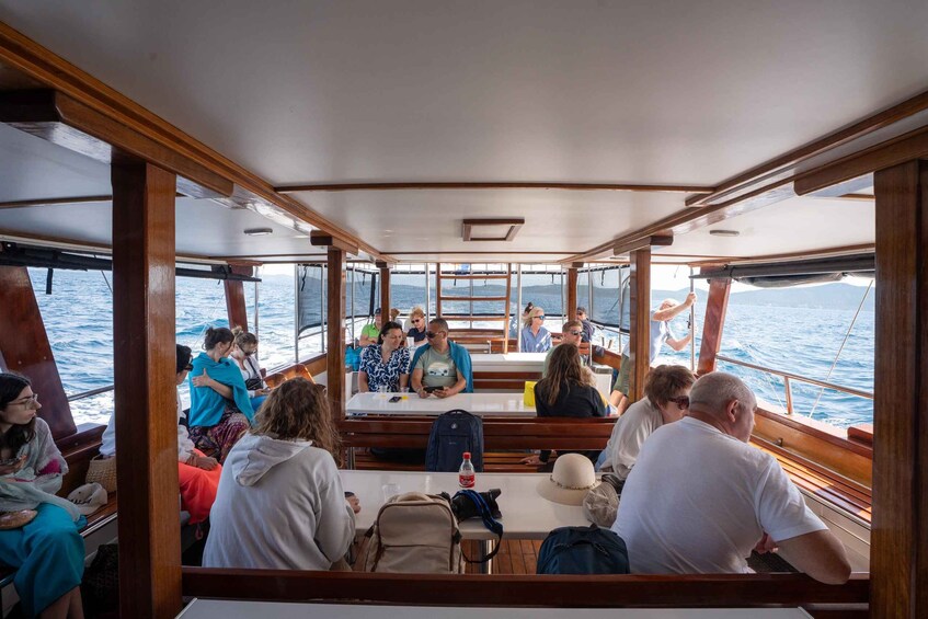 A group of people on a boat, some sitting and some standing, with a view of the sea and mountains in the background. 3
