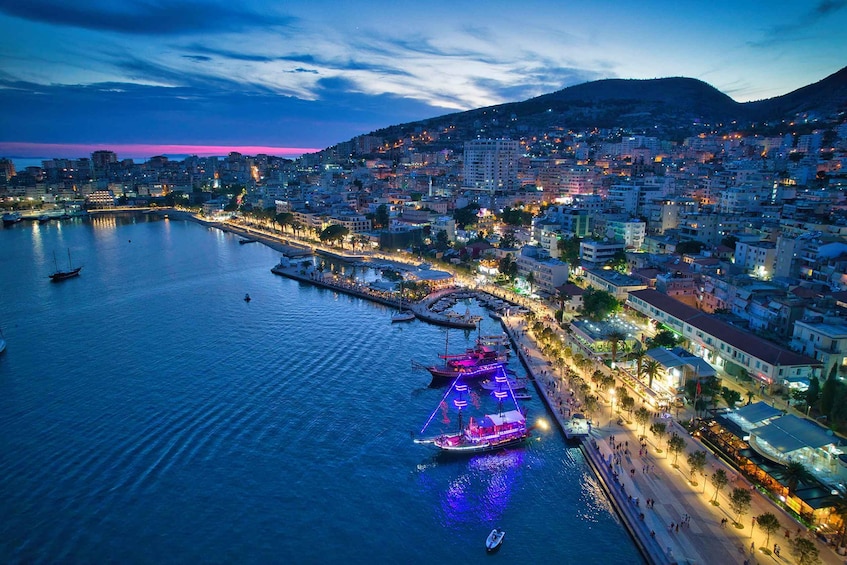 A coastal cityscape at dusk with illuminated boats docked at a marina, a promenade, and buildings along the waterfront. 5