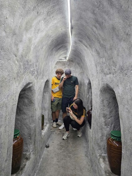 A narrow passageway with rough concrete walls, a person crouching and taking a photo, and two standing individuals. 5