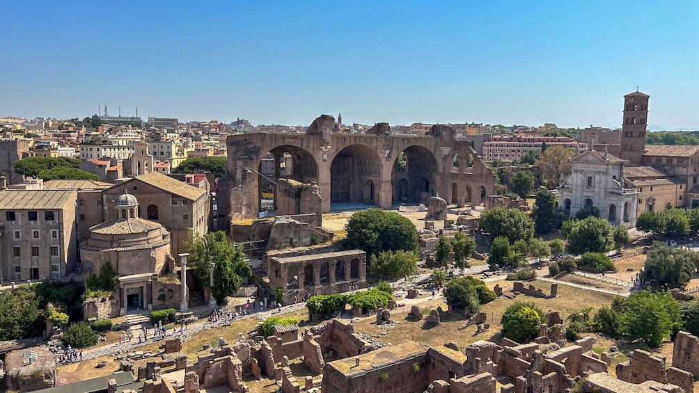 Ruinas antiguas con arcos y columnas, rodeadas de edificios modernos y un cielo despejado. 5