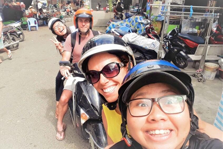 A group of people on motorcycles, some wearing helmets, in an outdoor setting with parked motorcycles and a market stall in the background. 3