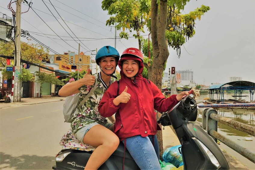 Two people on a scooter, one giving a thumbs up, with a river and buildings in the background. 2