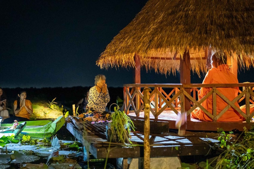 A thatched-roof structure with people sitting on a wooden platform by a body of water at night. 2