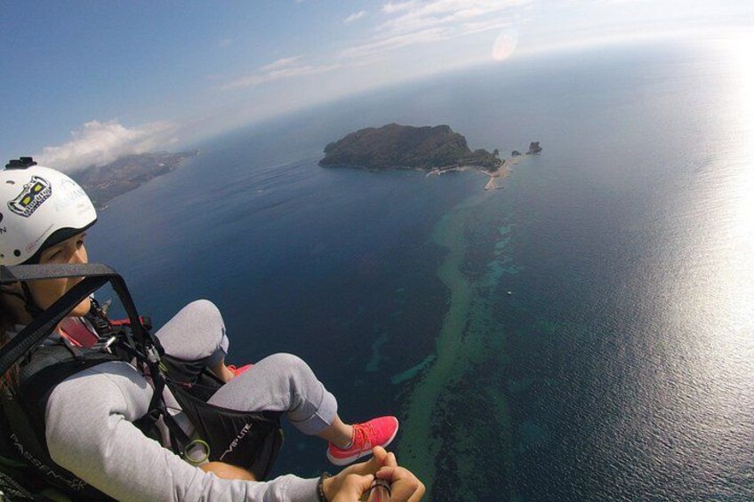 Una persona con arnés de paracaídas, casco y equipo de paracaídas, contemplando una zona costera con aguas azul turquesa y una pequeña isla. 2