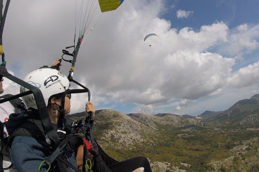 Una persona practicando parapente con casco y arnés, otra parapentando a lo lejos, y un paisaje montañoso. 4