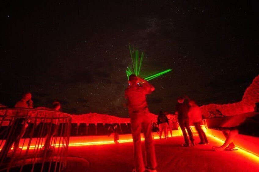 A nighttime scene with people standing on a platform under red lighting, with a green laser light in the center. 1