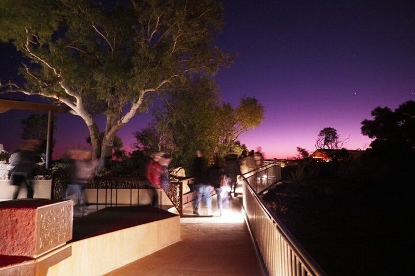 A nighttime walkway with people walking, a tree, and a purple sky. 3