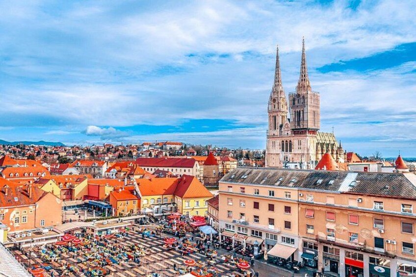 En stadsbild med en framträdande katedral med två torn, ett livligt torg med marknadsstånd och ett tätbebyggt område med byggnader som har röda tak. 3