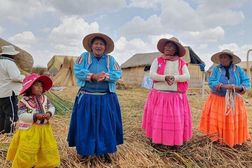 Four individuals in traditional attire standing in a rural setting with thatched-roof structures in the background. 4
