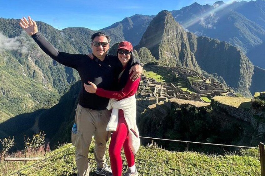 Two people standing in front of Machu Picchu with a mountain range in the background. 1