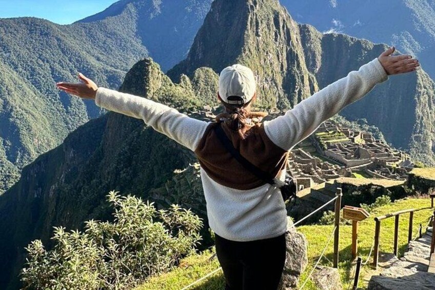 A person standing with arms outstretched, overlooking a mountainous landscape with a historical site below. 5