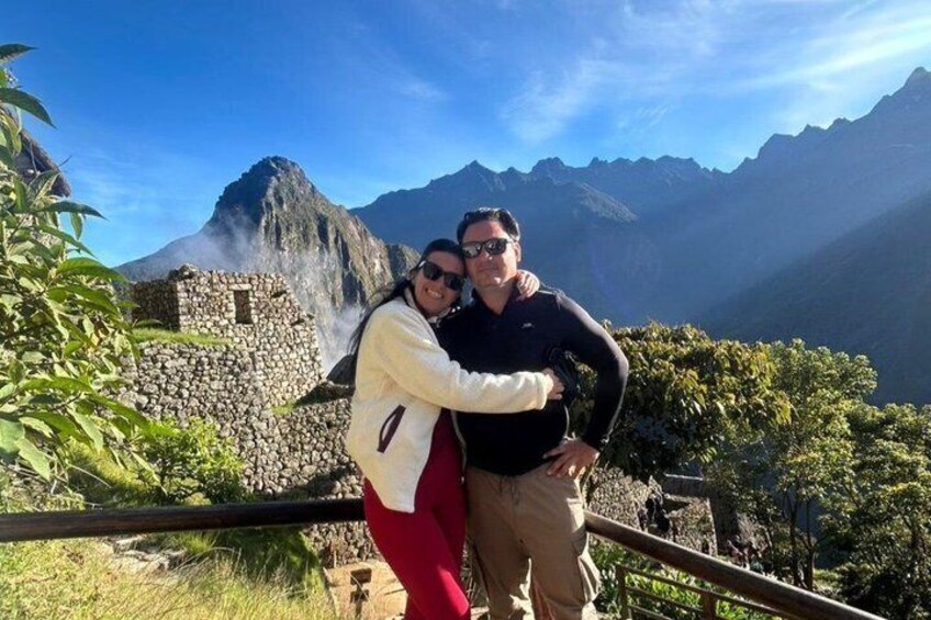 Two people standing by a stone structure with a mountainous backdrop. 1