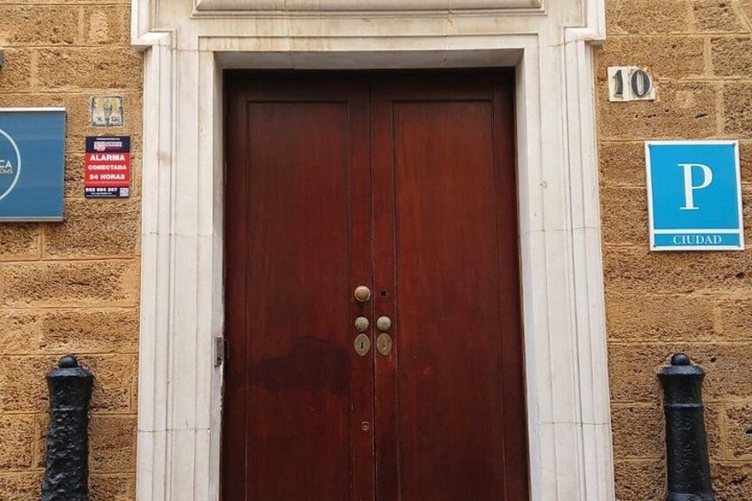 Una puerta doble de madera con un aldaban de latón, flanqueada por dos bolardos negros, y una señal de aparcamiento azul con la letra 'P' en la fachada de un edificio. 4