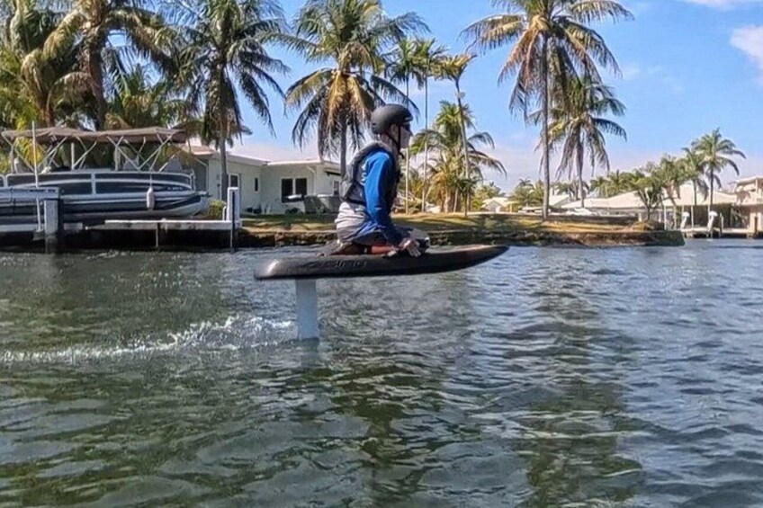 Une personne est debout sur une planche à pagaie, sur un plan d’eau, avec des palmiers et une maison en arrière-plan. 3
