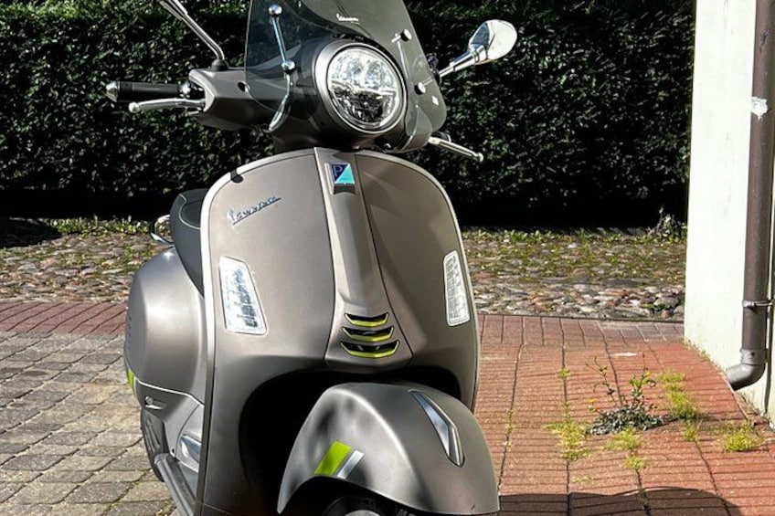 A grey Vespa scooter parked on a cobblestone path with a hedge and a building in the background. 3