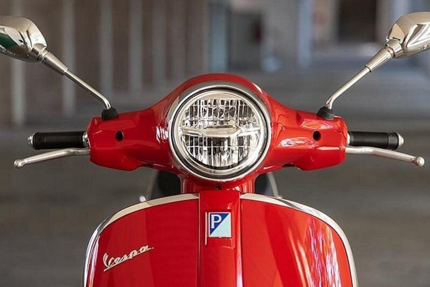 A red Vespa scooter with a round headlight and a blue parking sign. 1