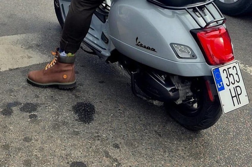 A silver Vespa scooter with a license plate, a person wearing brown boots, and a road with visible tire marks. 2
