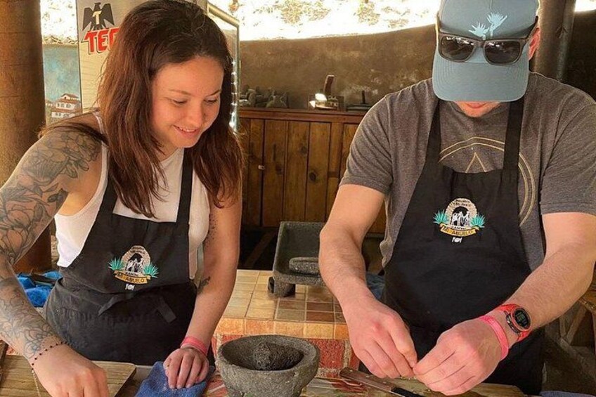 Twee personen met schorten, waarvan één een tatoeage heeft, die aan het werk zijn met een vijzel en stamper. 4