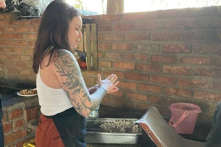Iemand met tatoeages op hun arm, gekleed in een witte top en rode broek, staat naast een bakstenen muur met een metalen dienblad en een roze bak. 5
