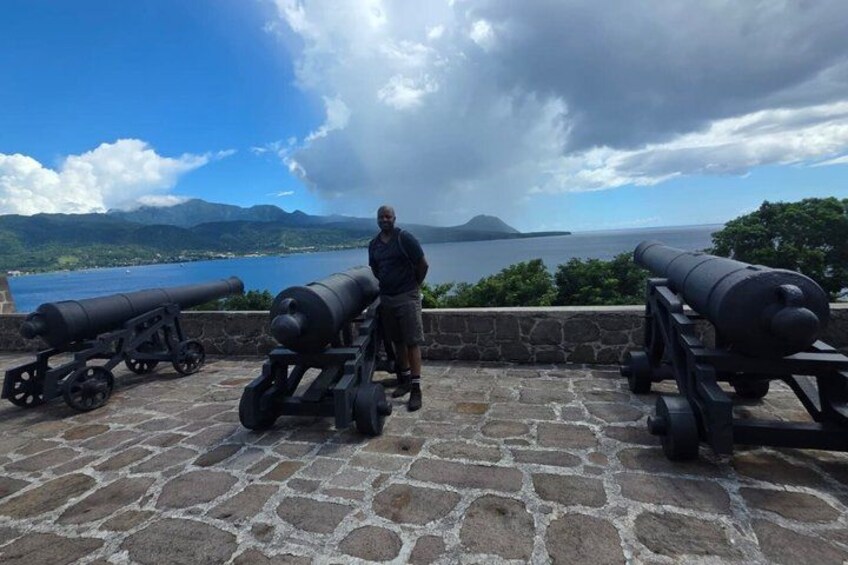 A person standing between two cannons on a stone platform overlooking a body of water and mountains. 3