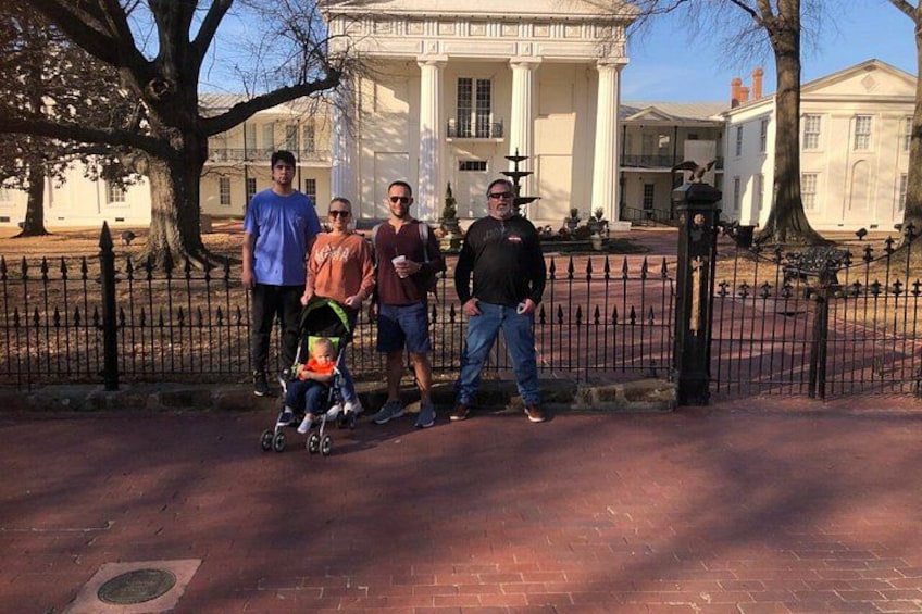 A group of people standing in front of a white building with columns, a brick pathway, and a metal fence. 2