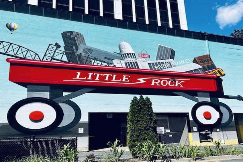 A red fire truck with 'LITTLE ROCK' and 'RIVER MARKET' on it, parked in front of a building with a blue facade. 3