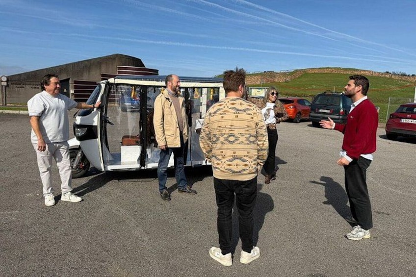 Un grupo de personas de pie alrededor de un vehículo pequeño y descubierto en un estacionamiento, con coches y un edificio al fondo. 1