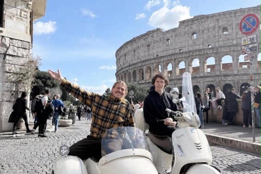 Two people on a scooter in front of the Colosseum with tourists in the background. 1