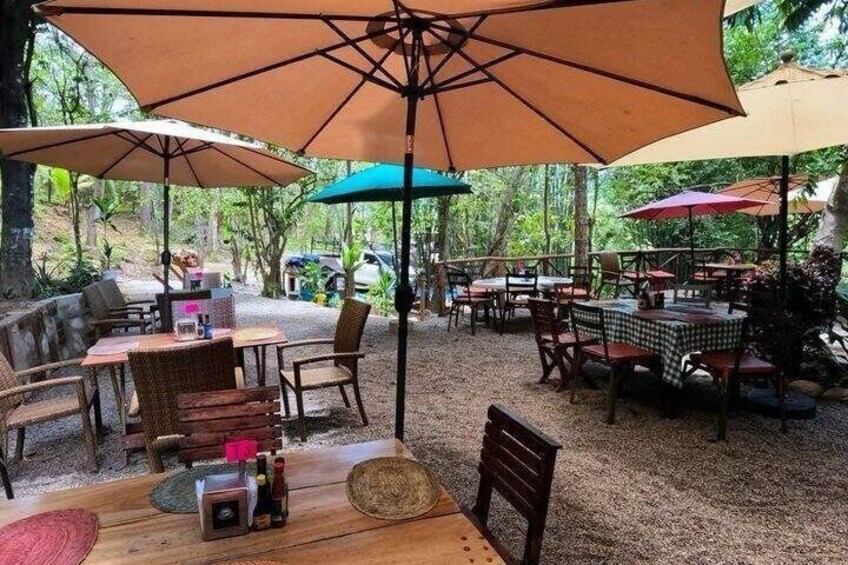 An outdoor café with tables and chairs under large umbrellas, surrounded by trees. 2