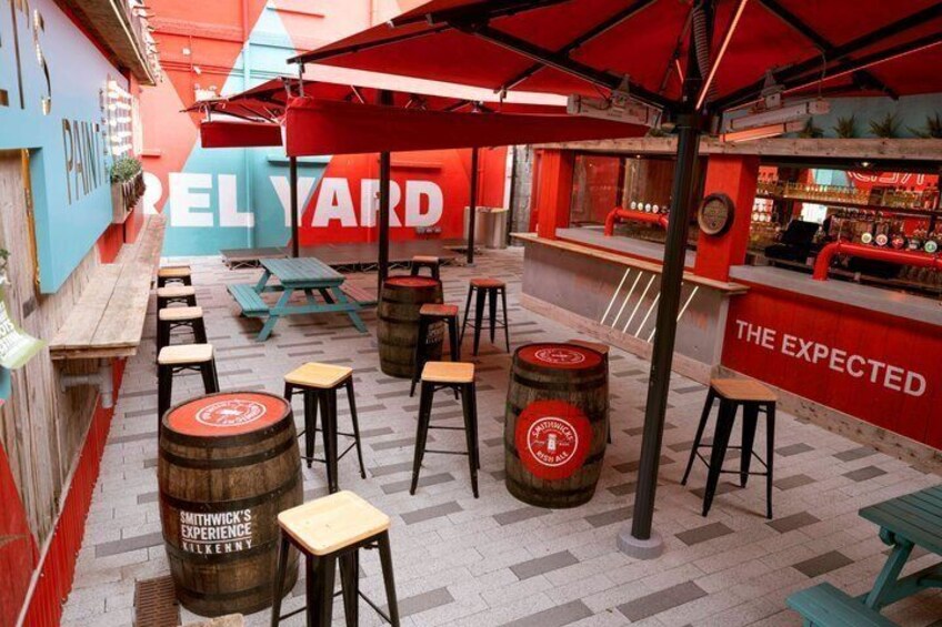 An outdoor bar area with wooden stools, red umbrellas, and barrels. 2