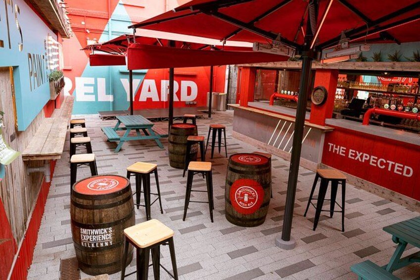 Een terras met houten krukken en vaten, een bar met een rood bord en een betegelde vloer. 3