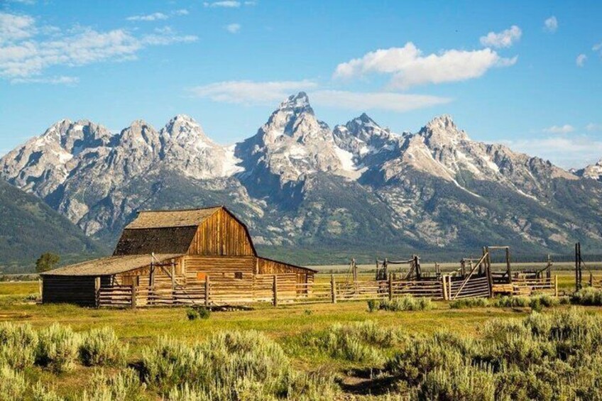 A wooden barn with a fenced area, surrounded by a grassy field and mountains in the background. 2