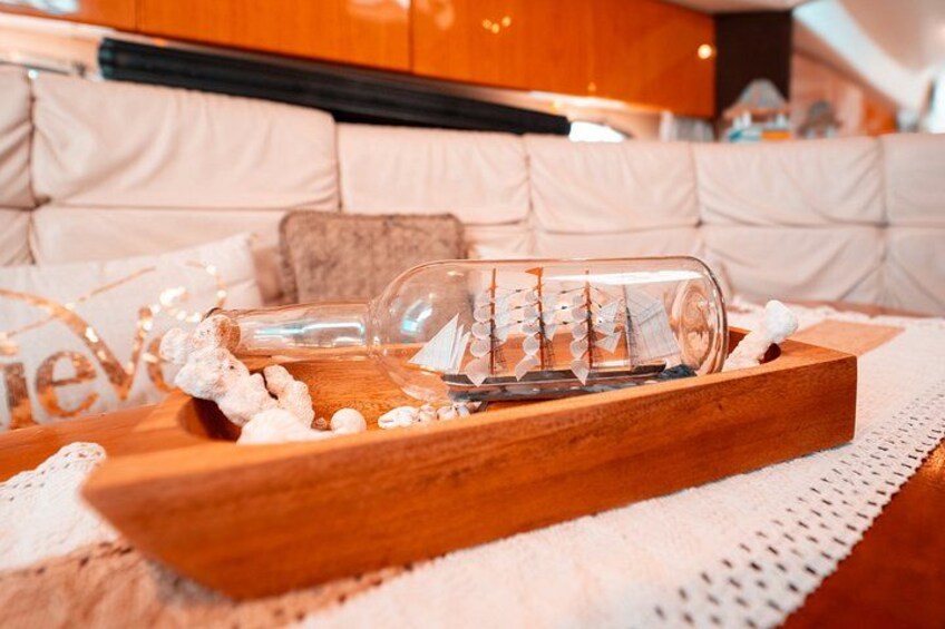 Una botella de vidrio con un modelo de barco en su interior, sobre una bandeja de madera con piedras decorativas, sobre una alfombra blanca texturizada. 4