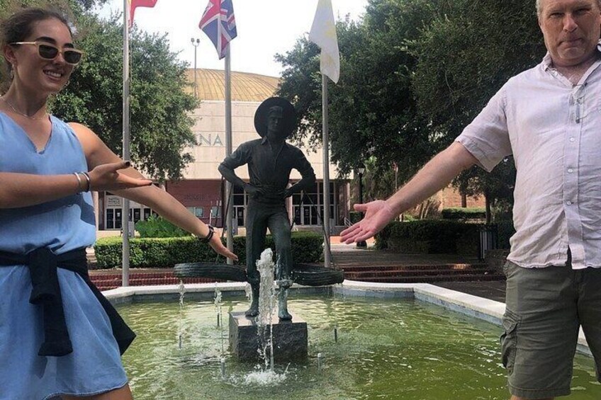 Ein Springbrunnen mit Wasserstrahlen, eine Statue und Flaggen in einer angelegten Grünfläche mit Bäumen und einem Gebäude im Hintergrund. 5