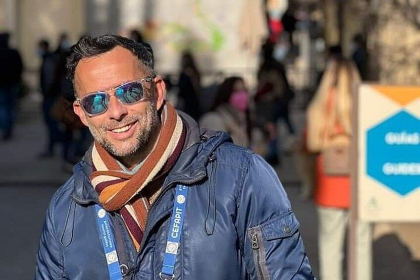 Eine Person mit blauer Jacke und Schal steht draußen, im Hintergrund verschwommene Gestalten und ein Schild. 1