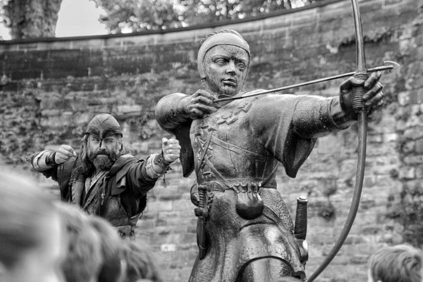 A medieval scene with a person in armor holding a bow, another in a hooded cloak, and a stone wall in the background. 2