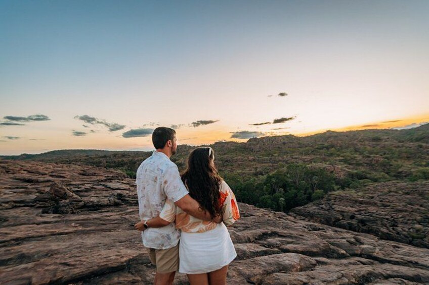 A couple standing on rocky terrain overlooking a hilly landscape during sunset. 1