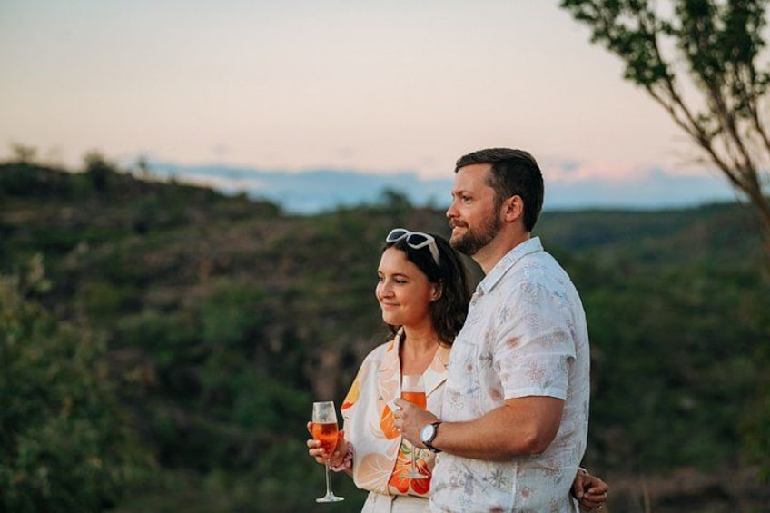 A couple standing outdoors with drinks, surrounded by greenery and hills. 3