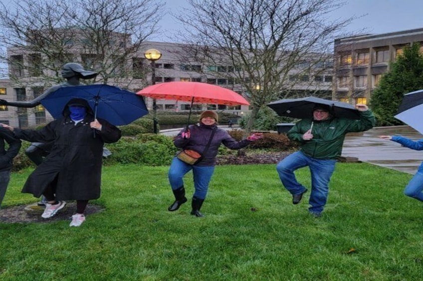 People dancing in a grassy area with umbrellas, a statue, and buildings in the background. 5