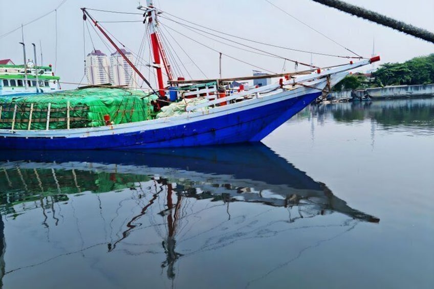 A blue fishing boat docked at a calm harbor with a green net on deck and a cityscape in the background. 2