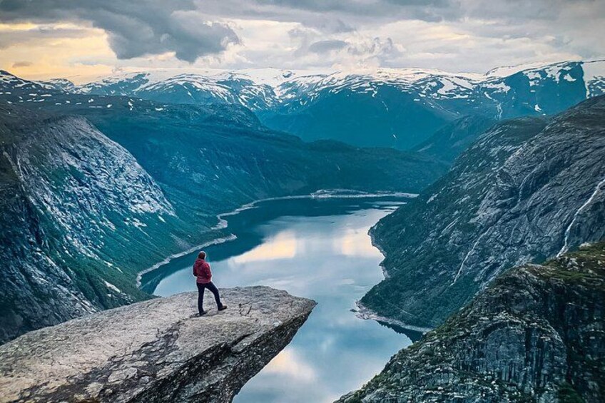 A person standing on a cliff overlooking a valley with a lake and mountains in the background. 1